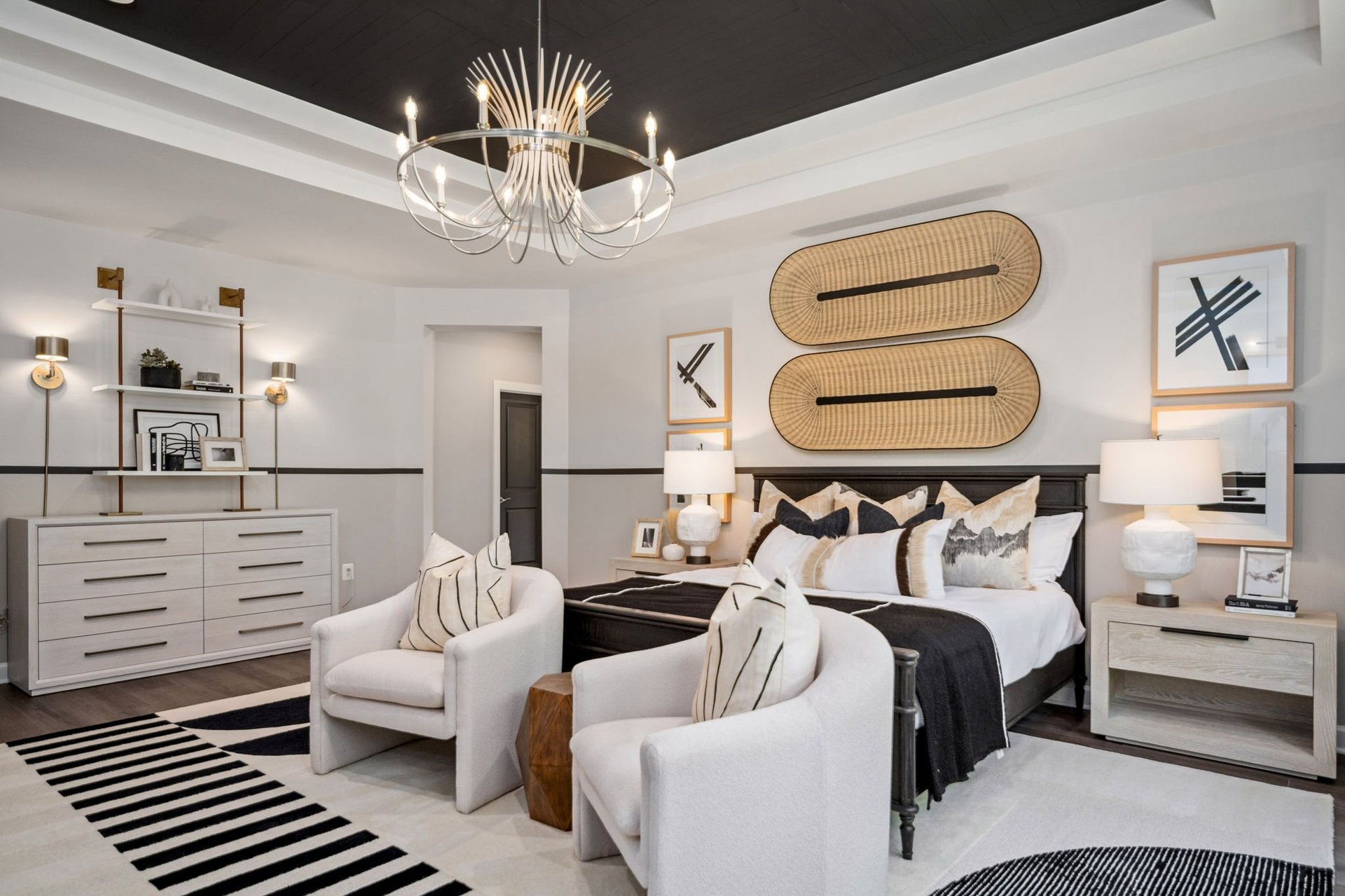 Modern bedroom with black and white color scheme, featuring a black bed with white bedding, two cream upholstered chairs, white dresser, and decorative woven wall panels
