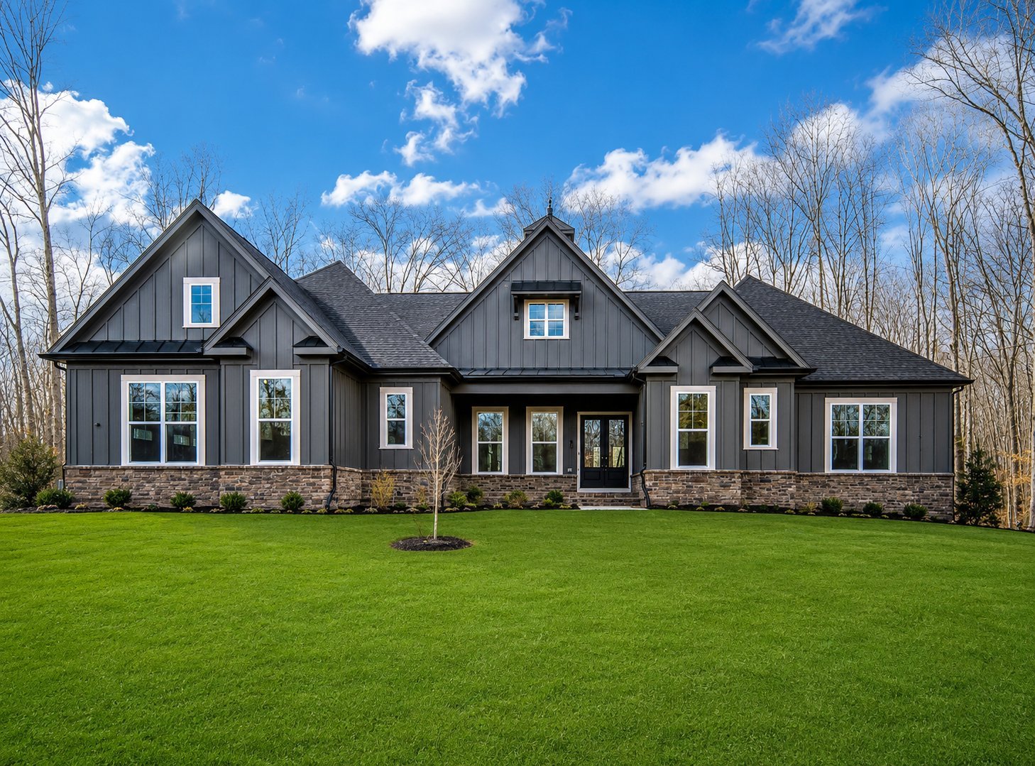 Modern two-story house with dark gray exterior, stone foundation, and multiple windows set on a large green lawn with bare trees in background